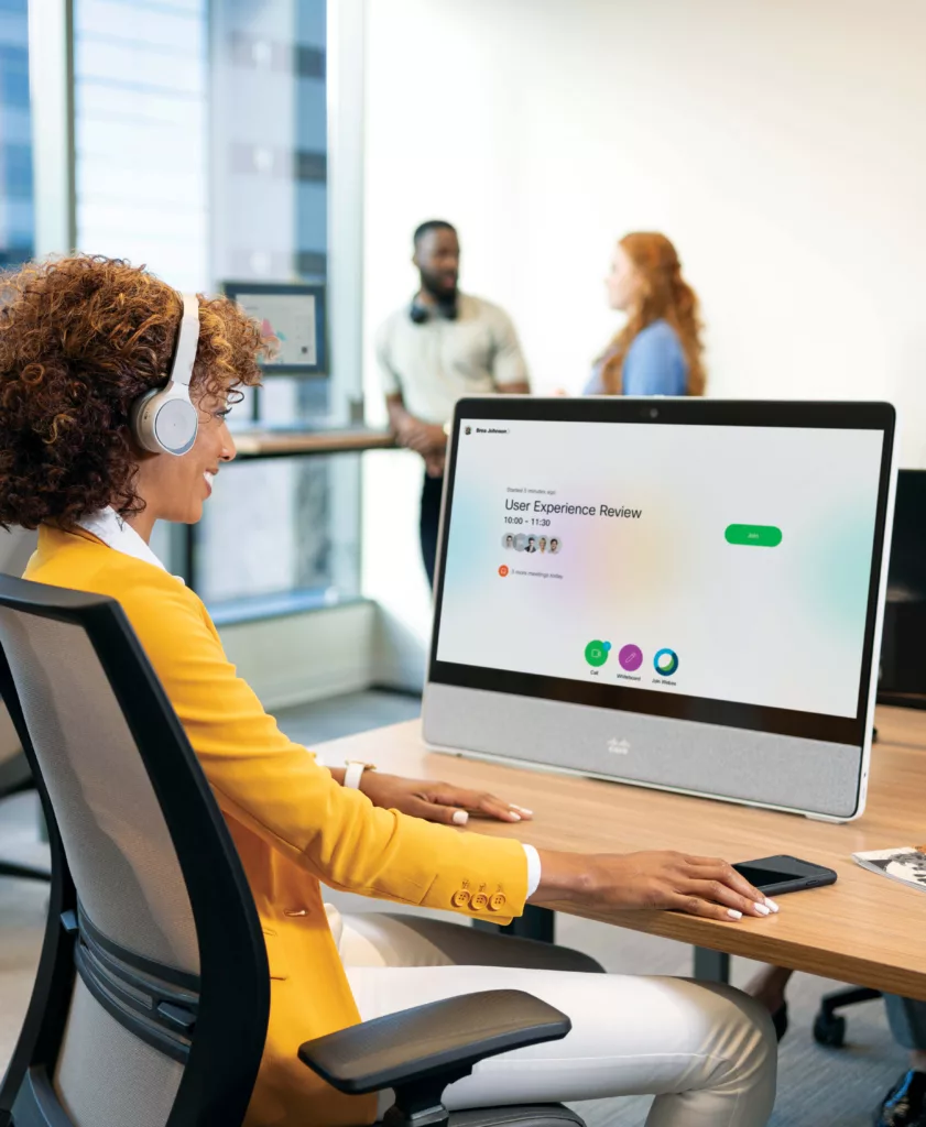 Woman sitting at a desk infront of the Cisco Webex Desk Pro and wearing Cisco bluetooth headphones getting ready to join a Webex meeting.