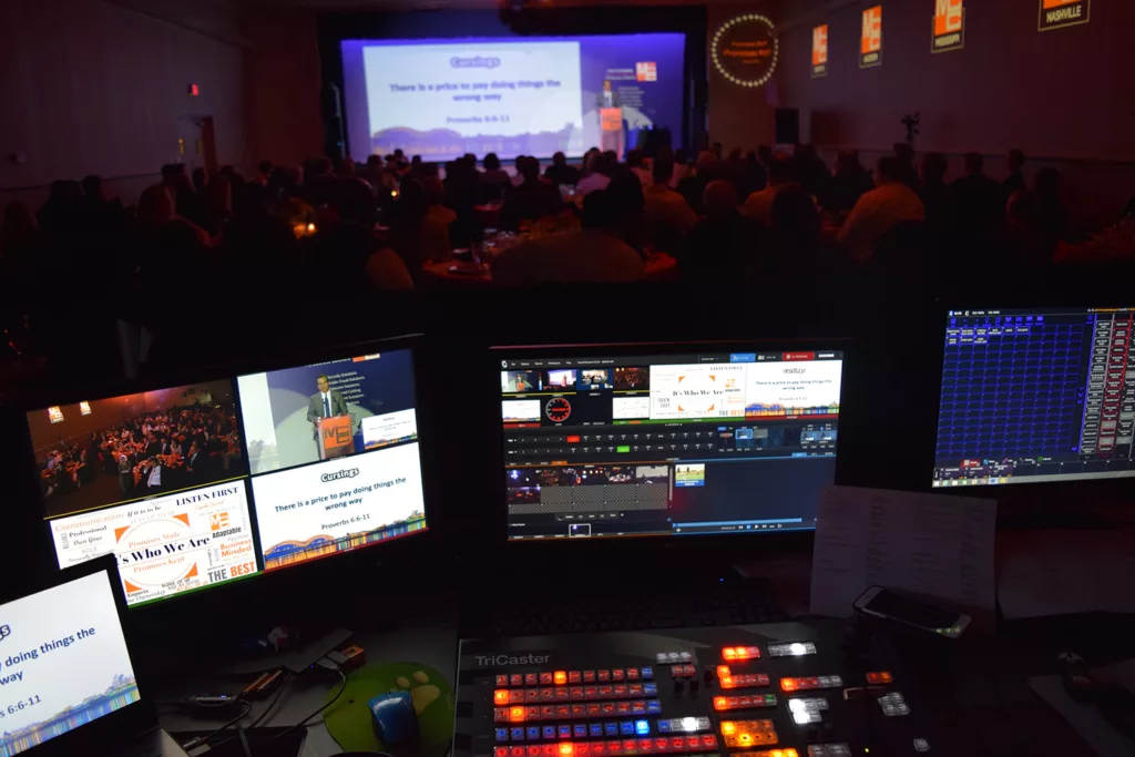 Image featuring computer screens in the foreground, displaying the NewTek Tricaster video production system in operation. In the blurred background, a large event is taking place, hinting at the real-world application of the advanced technology. This image showcases the seamless integration of high-tech video production into live events.