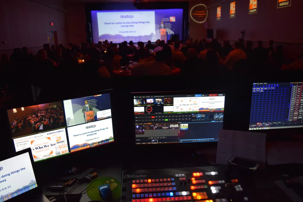 Image featuring computer screens in the foreground, displaying the newtek tricaster video production system in operation. In the blurred background, a large event is taking place, hinting at the real-world application of the advanced technology. This image showcases the seamless integration of high-tech video production into live events.